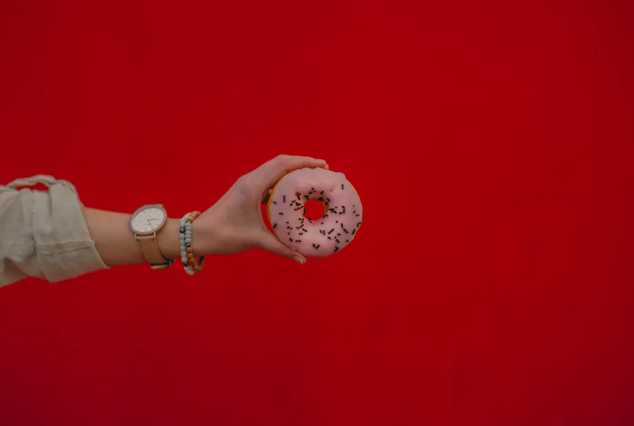 A hand holding a pink sprinkled donut against a flat red backdrop.