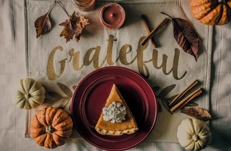 An autumn flat lay: a slice of pumpkin pie on a red plate, small pumpkins, cinnamon sticks and hand-lettered 'Grateful' on linen.
